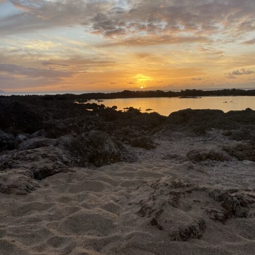 A sandy foreground with rocks, water and an orange sunset sky in the background